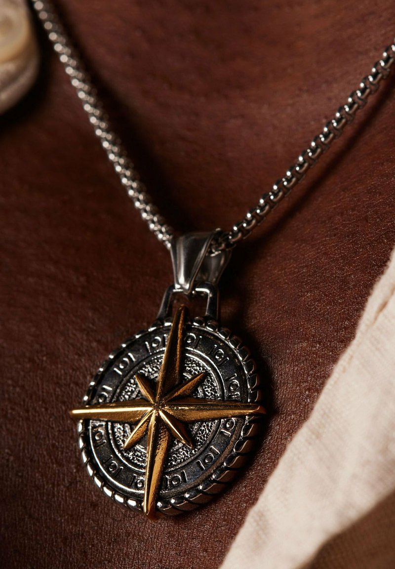 Zilveren ketting met een ronde hanger waarin een gouden kompasster is verwerkt, met een gestructureerde zilveren achtergrond en gegraveerde details rond de rand.