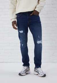 Jeans ajustados de color azul oscuro con detalles desgastados, que presentan parches rasgados. Combinados con zapatillas deportivas plateadas y un sudadera color crema.
