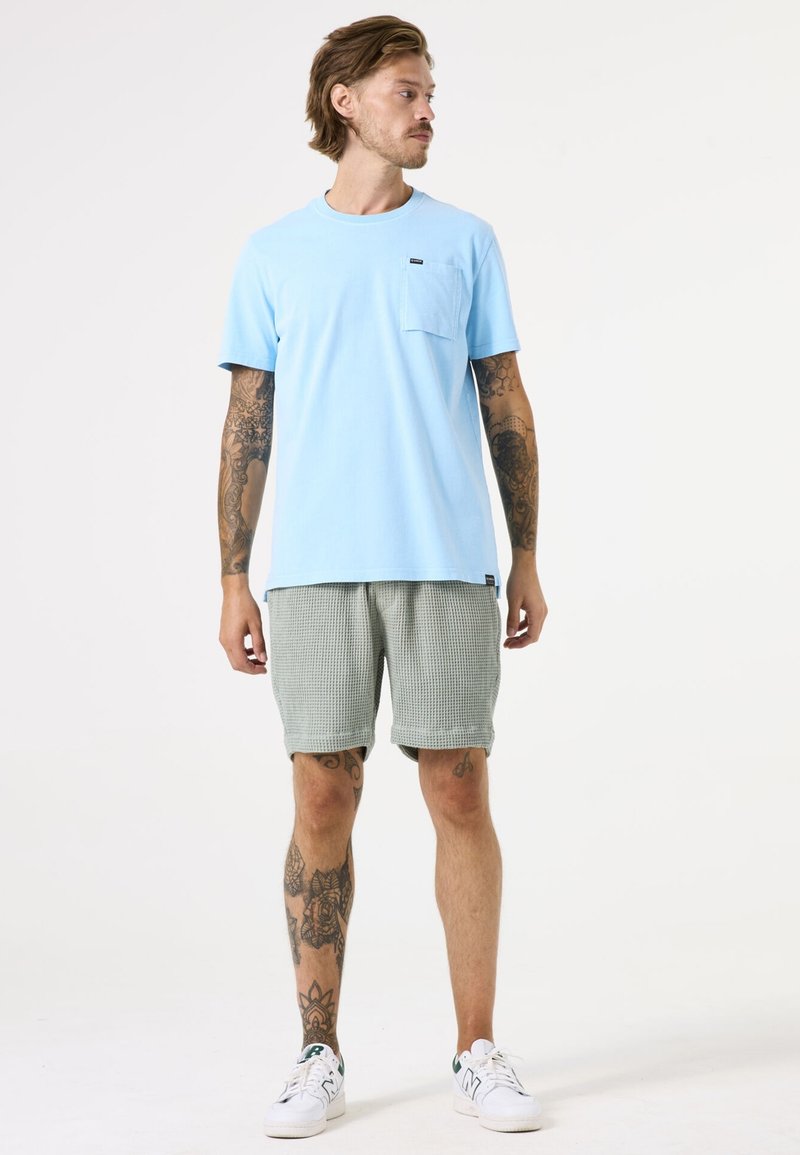 Lichtblauw katoenen T-shirt met een borstzak, gecombineerd met groene gestructureerde shorts en witte sneakers. Eenvoudig ontwerp, relaxte pasvorm.