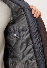 Fodera interna trapuntata nera di una giacca, con tasca con zip e texture liscia, abbinata a un materiale esterno marrone texturizzato.