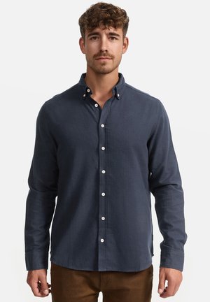 Homme aux cheveux bruns bouclés portant une chemise bleu marine à manches longues boutonnée et un pantalon marron, debout devant un fond blanc.