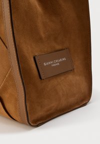 Καφέ τσάντα από σουέντ με ορθογώνια δερμάτινη ετικέτα που έχει ανάγλυφο το όνομα της μάρκας «Gianni Chiarini Firenze» και λεπτομέρειες με ραφές σε διαφορετικό χρώμα.