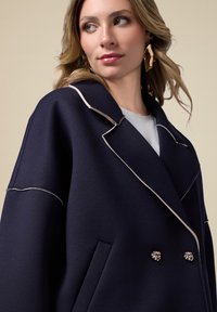 Cappotto doppiopetto navy con piping a contrasto chiaro, ampie rever e bottoni a nodo argentato ornamentali; presenta grandi tasche laterali e design strutturato.