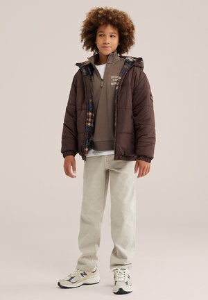 Junge mit lockigem Haar, der eine braune gesteppte Jacke über einem grauen Pullover mit halbem Reißverschluss und beigefarbene Hose mit weißen Sneakers trägt, steht und schaut seitlich.