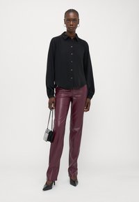 Camisa preta de manga longa com botão à frente, combinada com calças de pele sintética burgundy. Os acessórios incluem uma pequena mala preta e sapatos de salto agudo.