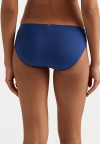 Bikini donji dio u mornarsko plavoj boji s glatkom teksturom, srednje visokim dizajnom i suptilnim zlatnim detaljem na stražnjem dijelu.