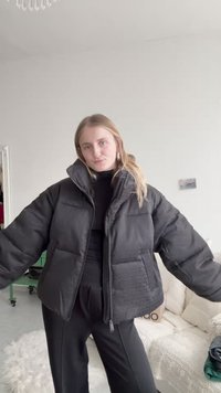 Crna pufasta jakna s teksturiranim uzorkom, širokog kroja, visokim ovratnikom, prednjim zatvaračem i dva bočna džepa. Nosena preko crne majice.