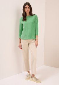 Eine Frau steht an einer beige Wand, trägt ein grünes Langarmoberteil, beige weite Hose und beige Turnschuhe und schaut in die Kamera.