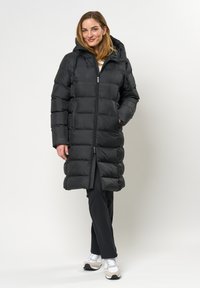 Svart pufferjakke med hette, quiltet design, kne-lengde, glidelås-lukking og sidelommer. Kombinert med svarte bukser og hvite joggesko.