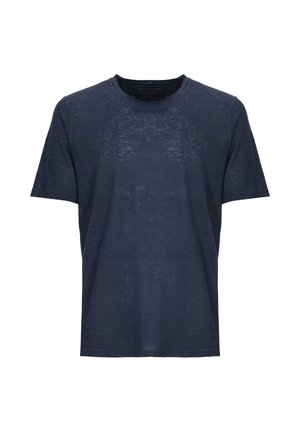 Uomo che indossa una maglietta blu navy trasparente, pantaloni bianchi con coulisse e sandali marroni, tiene in mano una camicia bianca a maniche lunghe con una mano in tasca.