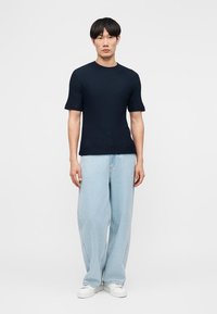 Νέος άνδρας που φοράει μια μπλε μπλούζα με κοντά μανίκια και υφή, ανοιχτό μπλε τζιν με φαρδιά πόδια και λευκά αθλητικά παπούτσια, στέκεται μπροστά σε ένα απλό φόντο.