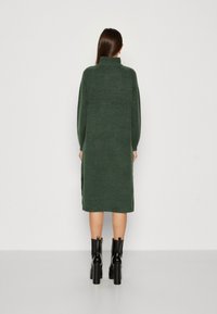 Donna in piedi di spalle, che indossa un lungo vestito a maglia verde scuro e stivali neri con tacco alto, su uno sfondo neutro.