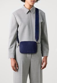 Borsa a tracolla in pelle blu con forma rettangolare, caratterizzata da una superficie testurizzata e una tracolla decorata, indossata sopra un outfit a quadretti.