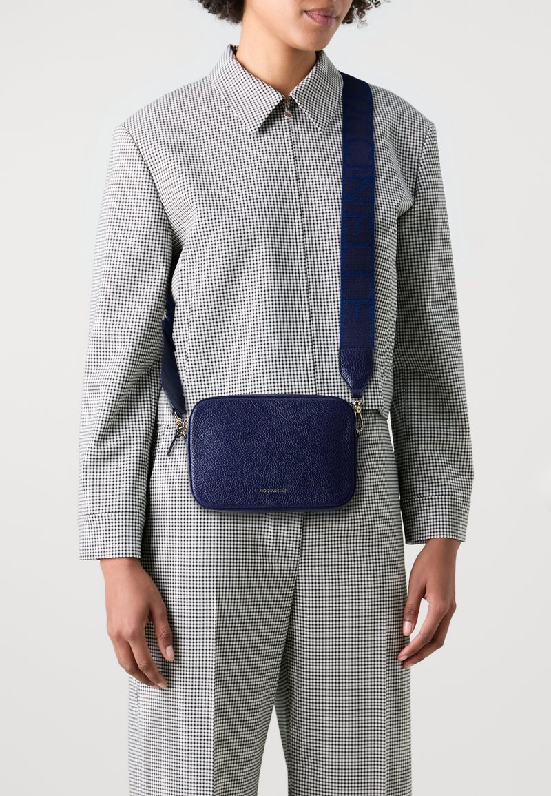 Borsa a tracolla in pelle blu con forma rettangolare, caratterizzata da una superficie testurizzata e una tracolla decorata, indossata sopra un outfit a quadretti.