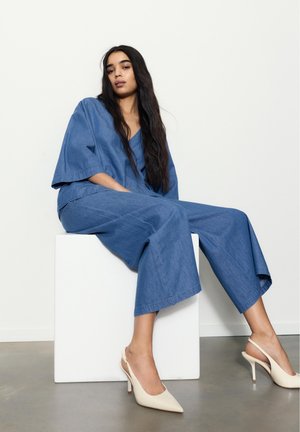 Tânără stând pe un cub alb, purtând o ținută largă din denim albastru și pantofi slingback crem cu toc înalt, pe un fundal simplu, deschis la culoare.