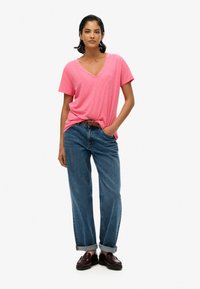 Camiseta de cuello en V rosa con mangas cortas, combinada con pantalones vaqueros azules holgados remangados en los puños, usados con zapatos burdeos.