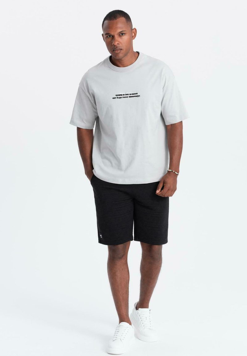 T-shirt grigia con testo nero, abbinata a pantaloni corti neri texturizzati. Sneakers bianche completano l'outfit. Design semplice e casual.