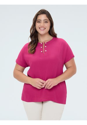 Femme souriante aux longs cheveux portant un haut rose vif à manches courtes avec des boutons en perle et un pantalon beige clair, debout devant un fond uni.
