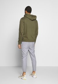 Olivgrön hoodie med snörning i huvan, som bärs med ljusgrå rutig byxor och krämfärgade sneakers. En enkel, avslappnad design med ribbade muddar.