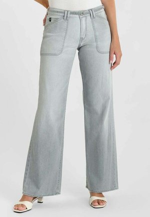 Vrouw draagt lichtgrijze jeans met wijde pijpen en voorzakken, met witte open-toe sandalen met hak, staand tegen een effen achtergrond.