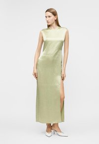 Γυναίκα με ίσια μαλλιά που φοράει αμάνικο ανοιχτό πράσινο σατέν φόρεμα με άνοιγμα στο πλάι, στέκεται και κοιτάζει προς τα πλάγια, φορώντας λευκές γόβες.