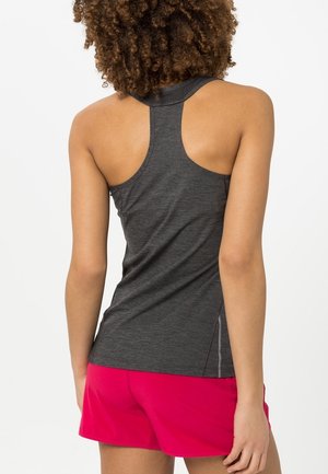 Žena s kudrnatými vlasy, oblečená v tmavě šedém tílku s průstřihy na zádech a jasně růžových šortkách, viděná zezadu na jednobarevném pozadí.