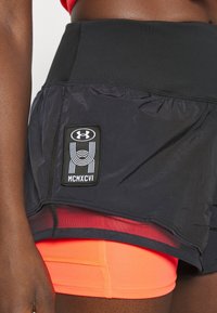 Svart träningsshorts med ett nätöverdrag, prydda med en Under Armour-logotyp. Klara orange underbyxor syns under.