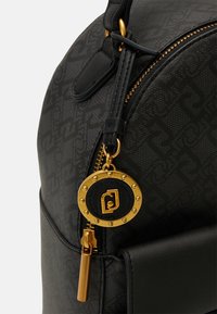 Gros plan d'un sac noir avec une fermeture éclair dorée et un pendentif rond doré et noir avec un logo stylisé et de petites clous décoratifs.
