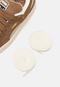 Brown suede sneaker with white accents and laces. Includes two rolled white shoelaces beside the shoe. Textured outsole visible.