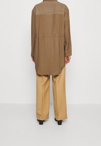 Hnědá košile bunda s zakulaceným lemem, dlouhými rukávy a detaily na zadním díle, ve spojení se světle béžovými širokými kalhotami a černými botami.