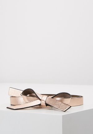 Ceinture fine en métal rose doré avec un détail nœud, posée à plat sur une surface blanche sur un fond clair uni.