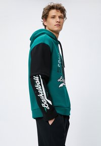 Sudadera con capucha en color teal y negro, con un gráfico en la parte frontal y texto blanco sobredimensionado en las mangas. Puños acanalados y bolsillo tipo canguro.