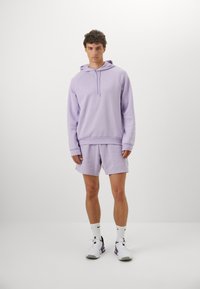 Lilla hettegenser og matchende shorts laget av myk stoff. Har en kengurulomme og ribbestrikkede mansjetter. Brukt med hvite joggesko.