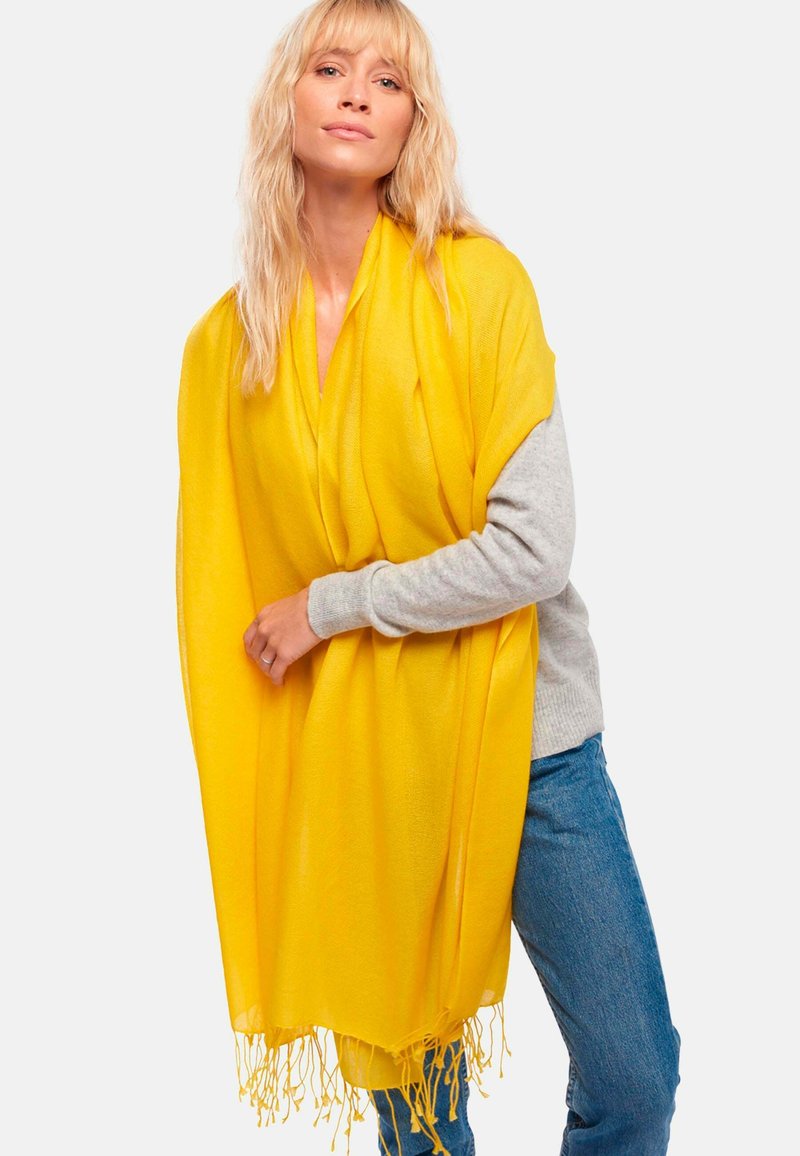 Châle jaune avec des bords à franges, fabriqué en tissu doux, porté sur un pull gris. Le châle tombe lâchement sur les épaules.