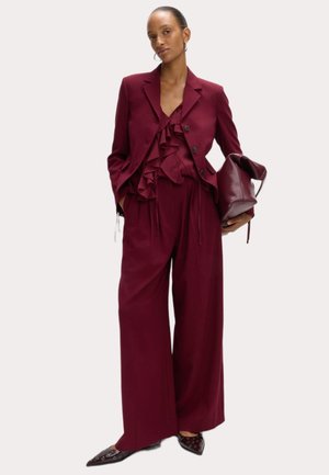 Costume bourgogne comprenant un chemisier à volants, un blazer ajusté avec des boutons, un pantalon à jambes larges et des chaussures à bout pointu avec des accents œillets.