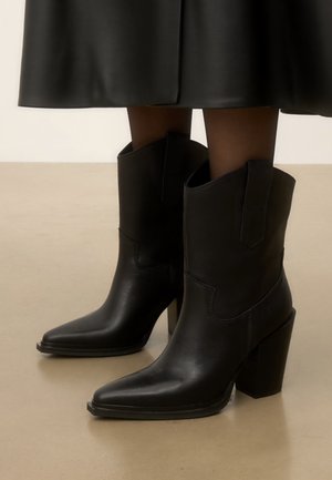 Bottes en cuir noir mi-mollet avec bouts pointus et talons blocs portées avec une jupe noire et des collants transparents sur un sol clair.