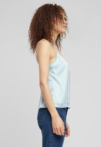 Camisolă din satin de culoare albastru deschis, cu bretele subțiri, o croială lejeră și o textură netedă. Haina coboară puțin sub talie.