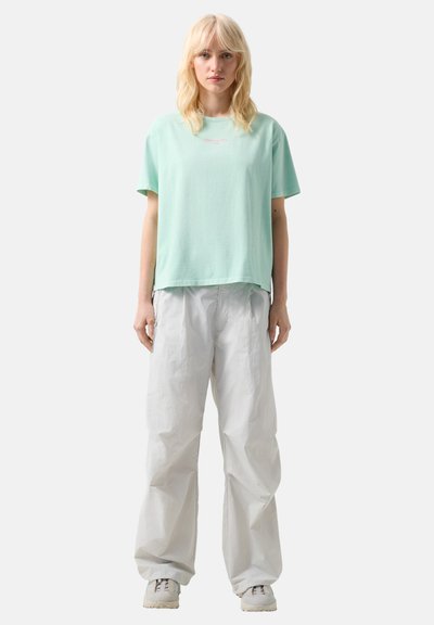 Blonde vrouw die een oversized mintgroen T-shirt draagt, losse witte broek en lichtgrijze sneakers, staand tegen een effen achtergrond.