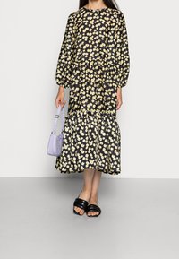 Schwarzes Blumenkleid mit gelben Blumen, Ballonärmeln und einem gestuften Saum. Eine kleine Handtasche aus Lavendel und schwarze Schlappen runden den Look ab.