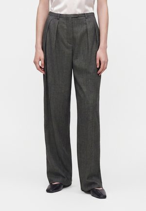 Femme vêtue d’un pantalon gris à carreaux taille haute, à jambes larges avec des plis sur le devant et des chaussures plates noires, debout devant un fond uni.