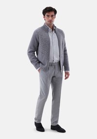 Mann steht und trägt eine graue Strickjacke mit Reißverschluss, ein hellgraues Hemd mit Knöpfen, graue Hose und schwarze Slipper vor weißem Hintergrund.