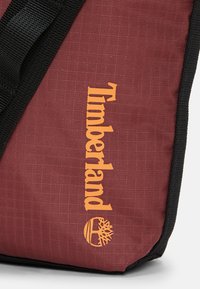 Borsa in tessuto rossa con dettagli neri, caratterizzata dal logo "Timberland" arancione e un emblema con un design ad albero. Tessuto morbido, spallacci robusti.