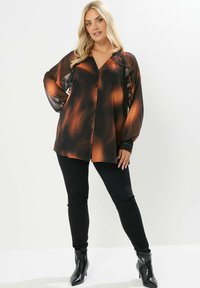 Camisa de manga larga abotonada en degradado naranja y negro con detalles con volantes, combinada con pantalones negros slim-fit y botines de punta. 
