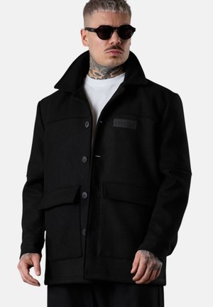 Mann mit Hals- und Handtattoos trägt schwarzen, geknöpften Blazer über weißem Hemd und dunkler Sonnenbrille vor hellem, einfarbigem Hintergrund.