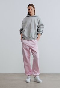 Νεαρό άτομο που στέκεται φορώντας γκρι φούτερ με κουκούλα, ανοιχτοροζ φόρμα και λευκά αθλητικά παπούτσια, μπροστά σε ένα απλό λευκό φόντο.