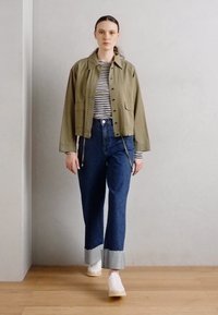 Veste raccourcie olive avec fermeture à boutons sur une chemise à rayures noires et blanches, associée à un jean bleu foncé taille haute et des chaussures blanches.