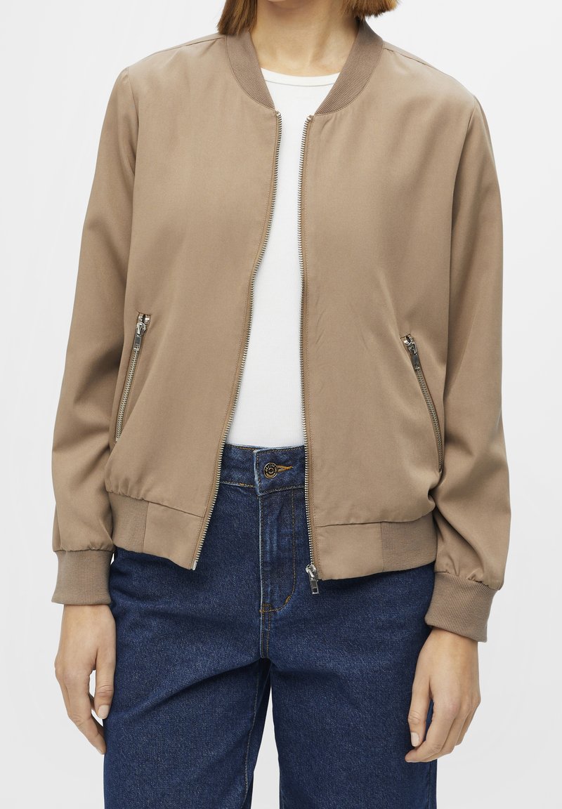 Tanfarbener Bomberjacke mit Reißverschluss, seitlichen Taschen, gerippten Bündchen und Saum. Trägt sie über einem weißen Oberteil, kombiniert mit dunkelblauen Jeans.