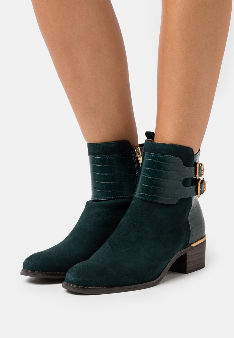 Bottes hauteur cheville en daim vert foncé avec des lanières en cuir texturé, des boucles dorées, des fermetures éclair latérales et des talons blocs bas portées sur des jambes nues.