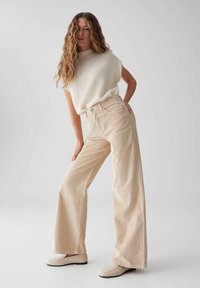 Beige Cordhosen mit weitem Bein und hoher Taille, kombiniert mit einem weißen, flauschigen, ärmellosen Oberteil. Das Model steht in einer entspannten Pose.