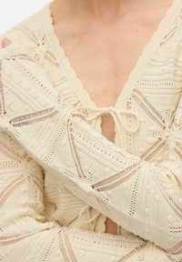 Gebreide semi-doorzichtige crèmekleurige top met een V-hals, strikdetail, gestructureerde patronen en ingewikkelde opengewerkte ontwerpen.